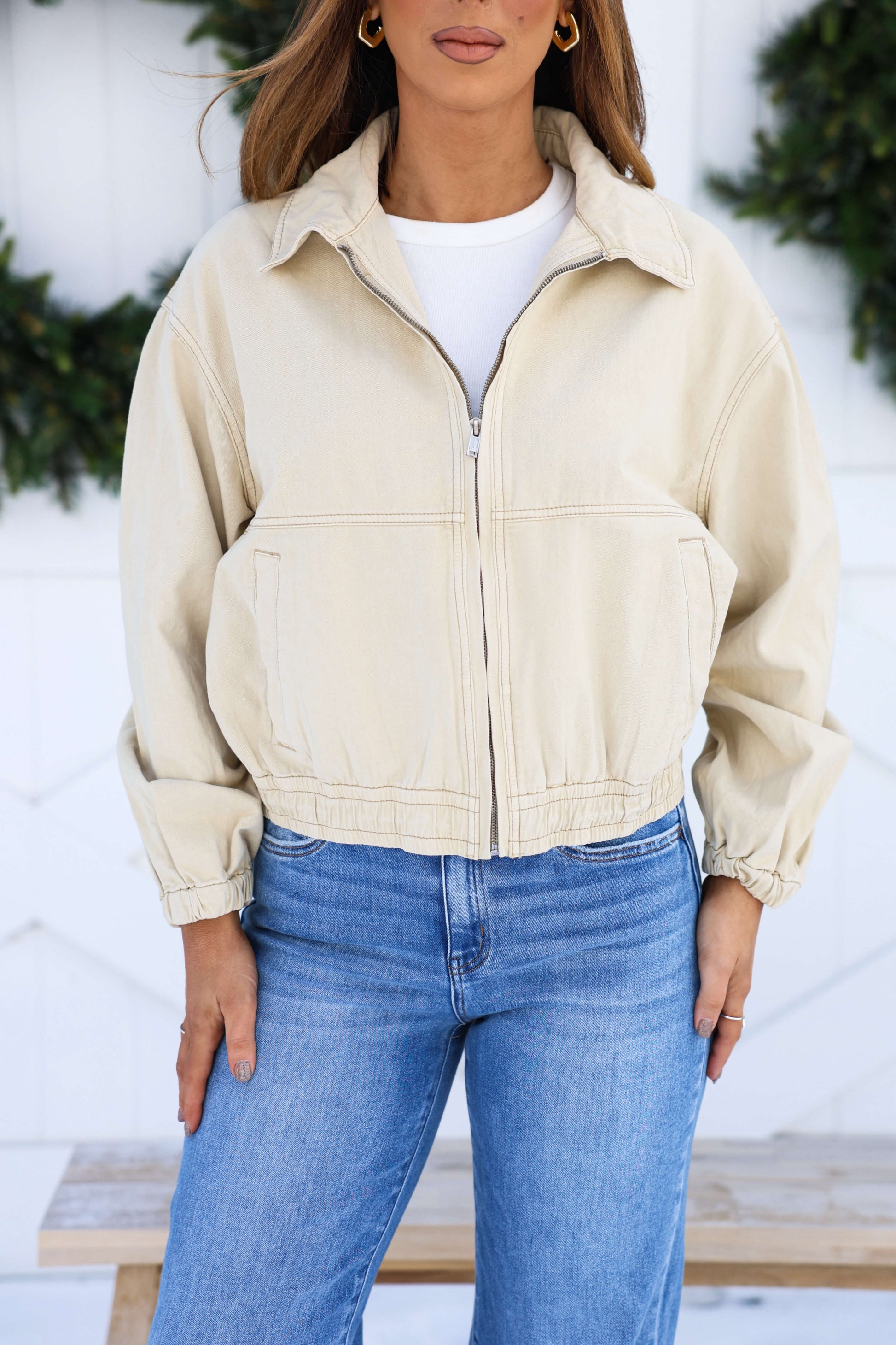 A woman in a Free People Cream Birdie Jacket over a white shirt and blue jeans stands outdoors before greenery.
