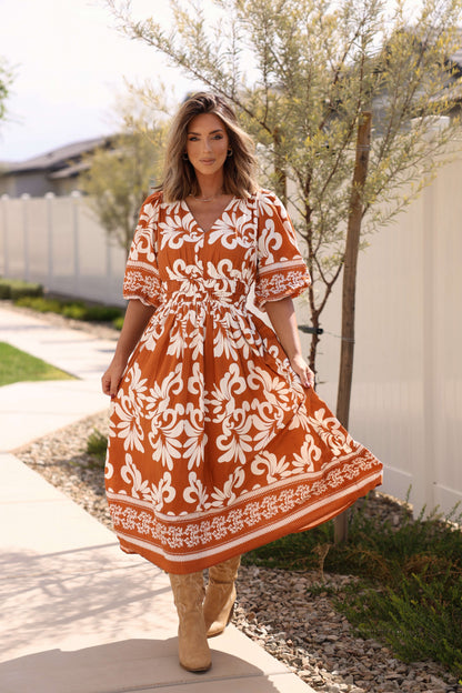 A woman in the Cider Lane Caramel Print Midi Dress stands on a sidewalk, holding her skirt with trees and a white fence behind her.