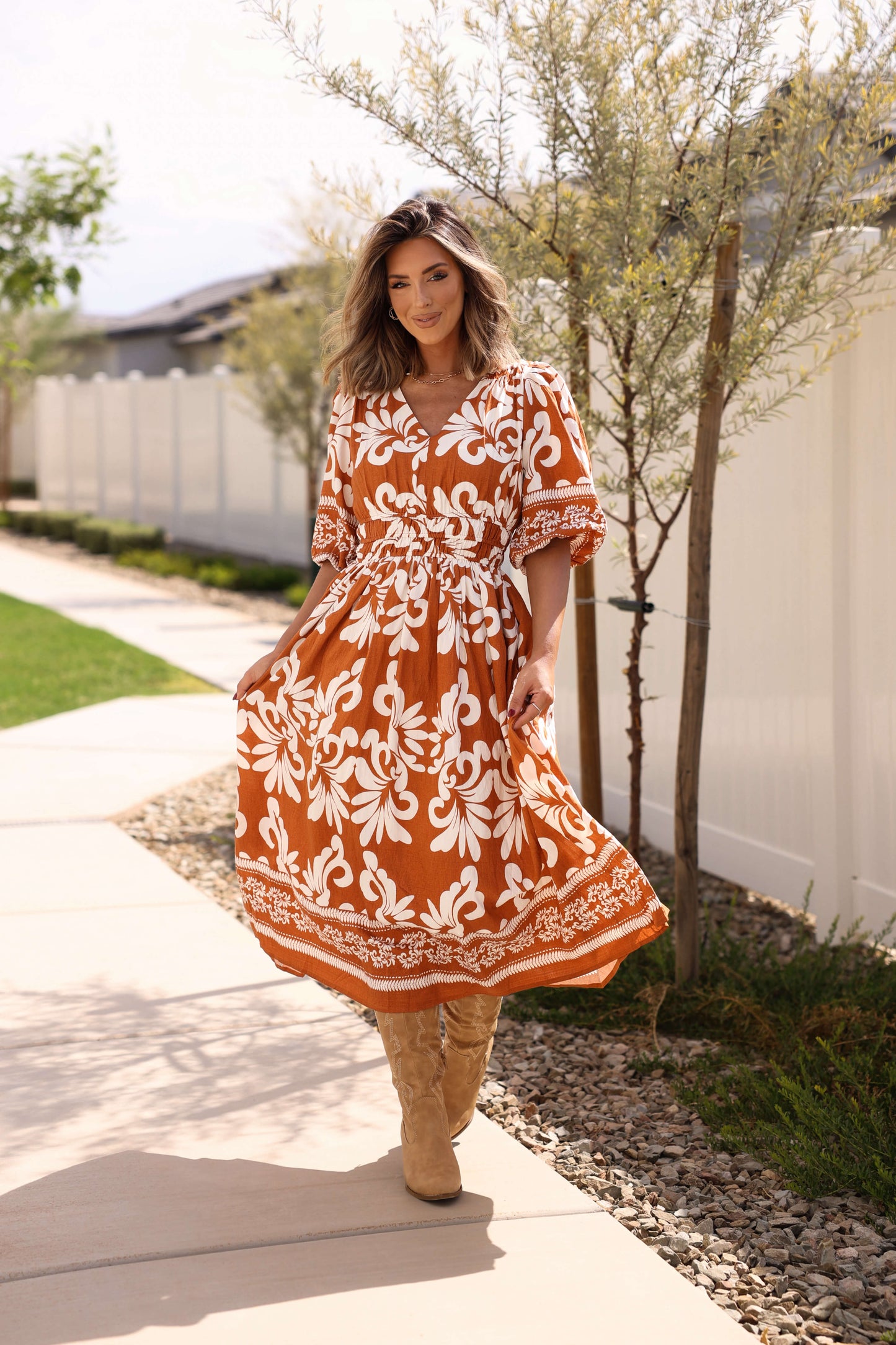 Woman in the Cider Lane Caramel Print Midi Dress walks on a sidewalk by a white fence and small trees.