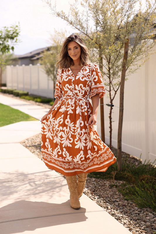 Woman in the Cider Lane Caramel Print Midi Dress walks on a sidewalk by a white fence and small trees.