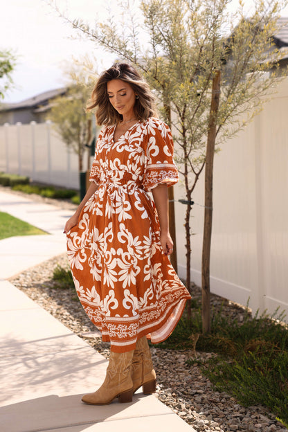 Woman in a Cider Lane Caramel Print Midi Dress stands on a sunny sidewalk by a white fence and trees.