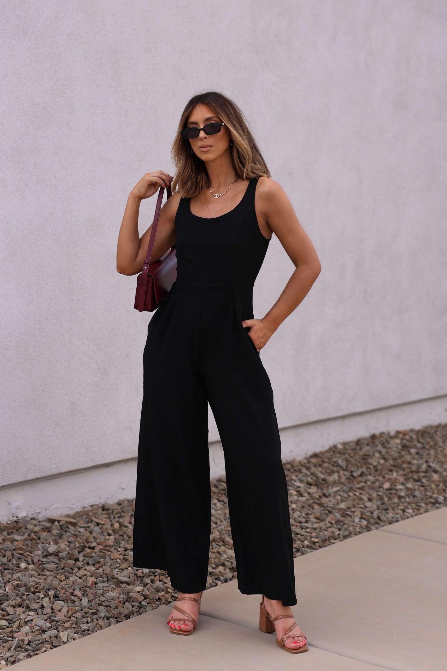 Woman wears the Hunter and Brown Black Pleated Jumpsuit with a scoop neckline and wide-leg fit, standing confidently outdoors.