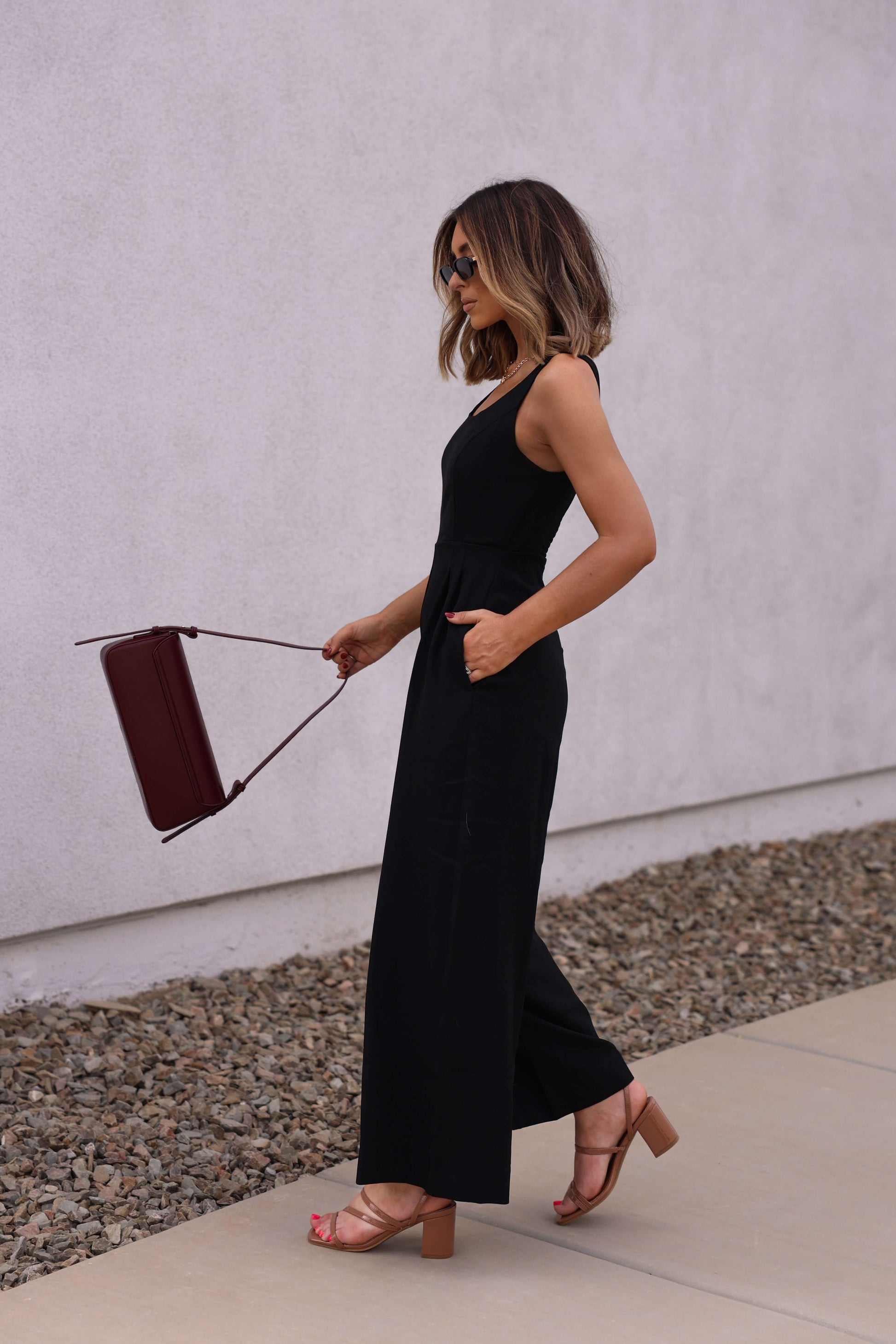 Woman wears the Hunter and Brown Black Pleated Jumpsuit while walking outdoors, carrying a maroon handbag by a white wall.
