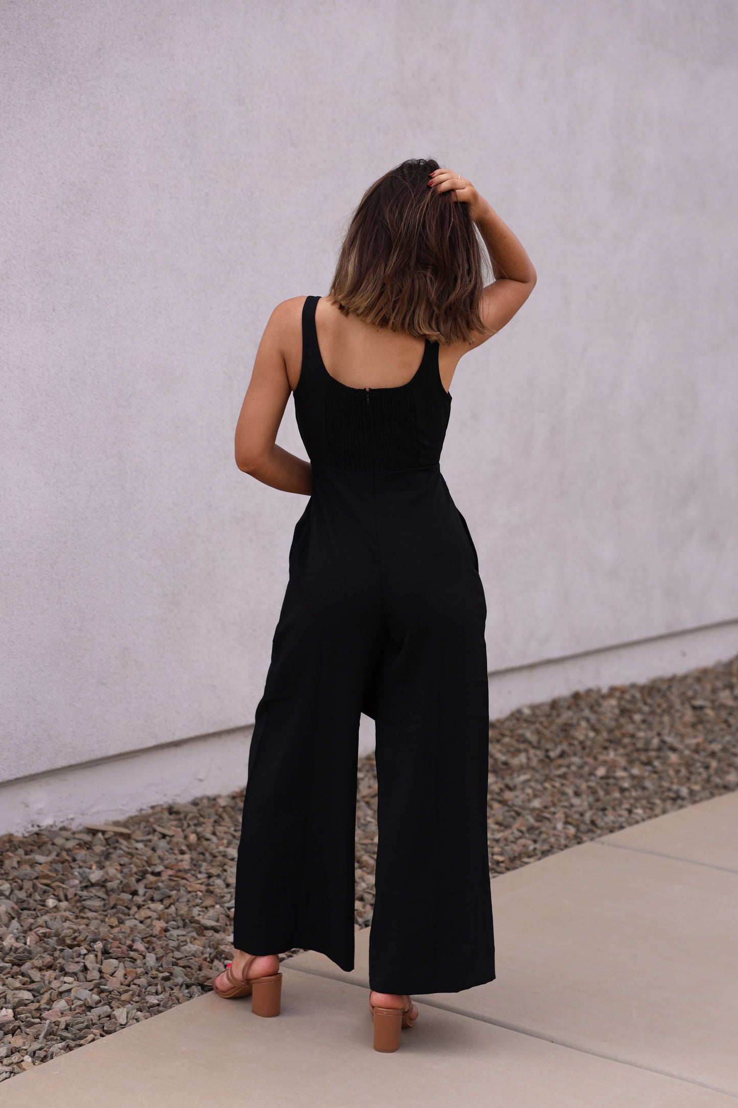 A woman wears the Hunter and Brown Black Pleated Jumpsuit, a sleeveless wide-leg style, standing outdoors and touching her hair.