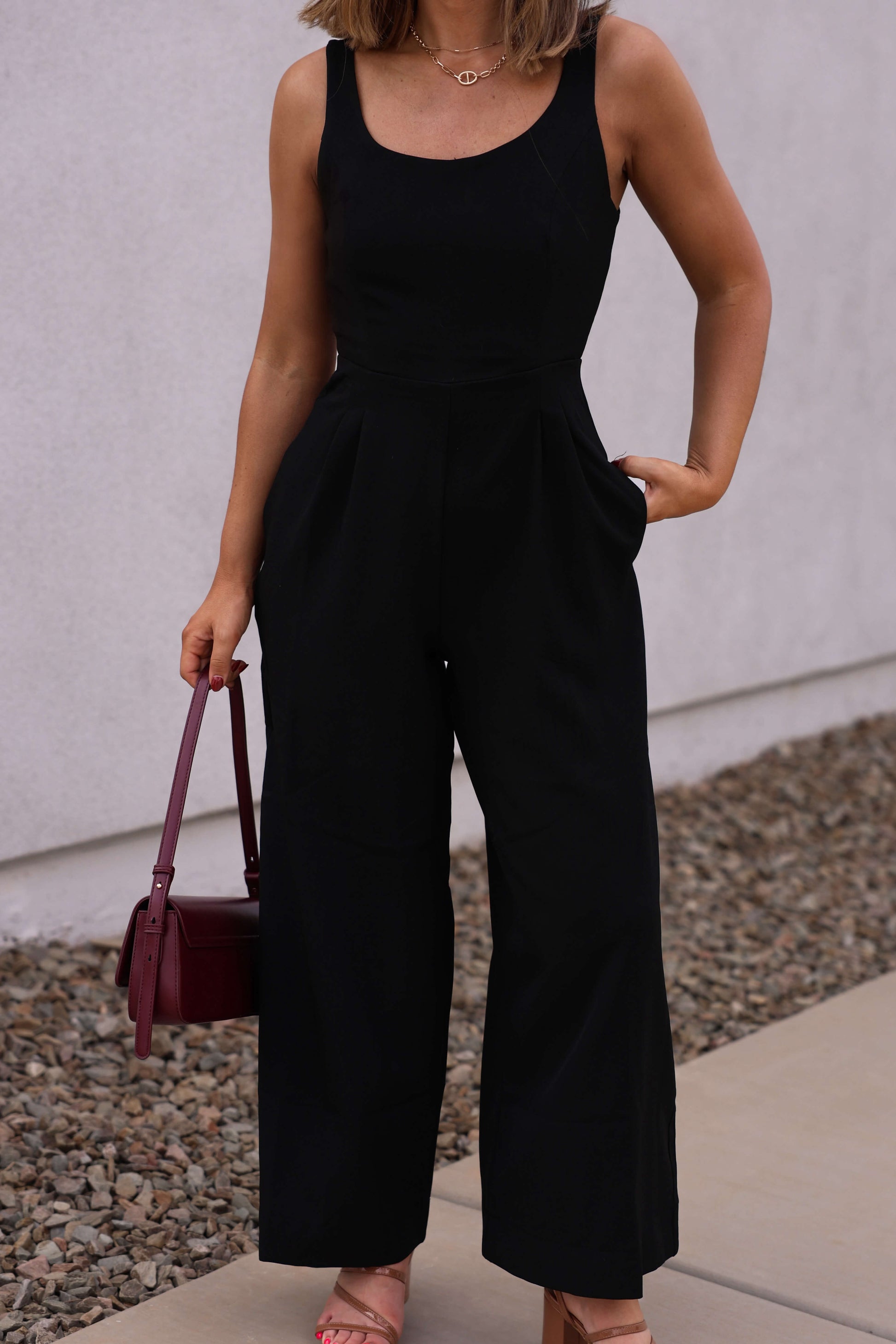 Woman wearing the Hunter and Brown Black Pleated Jumpsuit, holding a burgundy handbag on a rocky sidewalk.