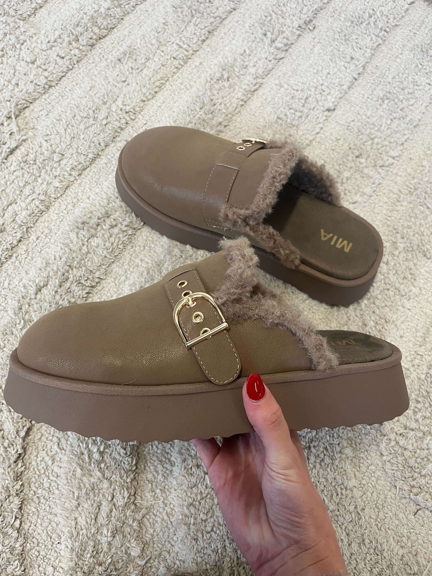A hand holds the MIA Sugary Taupe Sherpa Mule with gold buckle; matching slipper and textured rug in the background.