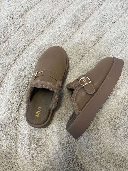 A pair of MIA Sugary Taupe Sherpa Mule clogs with fur lining and gold buckles on a textured beige carpet.