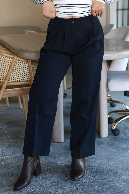 A person in Anabelle Black Pleated Trousers, brown heeled boots, and a striped top stands near a dining table and chairs.