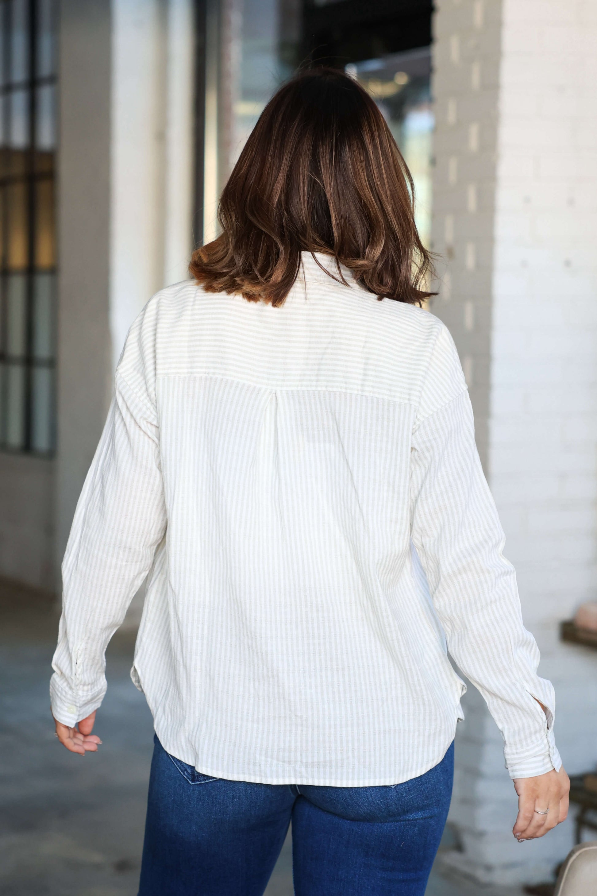 A person with shoulder-length brown hair wears the Thread and Supply Edison Tan Striped Shirt and blue jeans, facing away indoors.