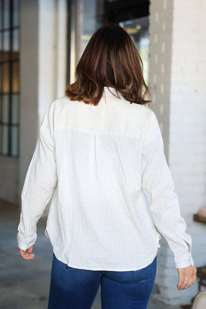 A person with shoulder-length brown hair wears the Thread and Supply Edison Tan Striped Shirt and blue jeans, facing away indoors.
