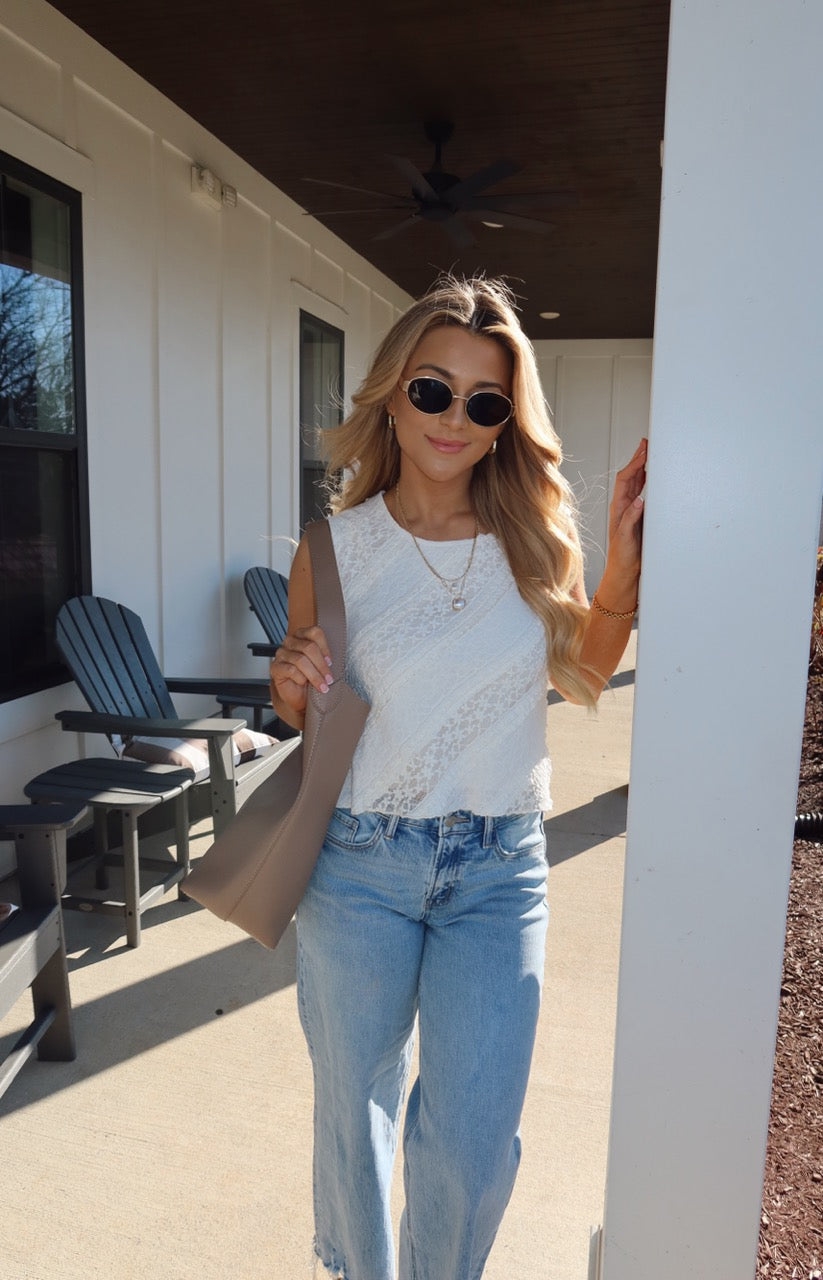 Woman with long blonde hair in sunglasses and blue jeans wears the Mika Ivory Ruffled Lace Top and holds a taupe bag on a sunny porch.