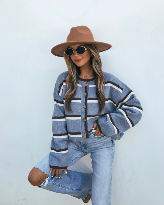 A person wears an oversized Striped Sweater - Denim Blue, light-wash distressed jeans, a wide-brimmed hat, and sunglasses.