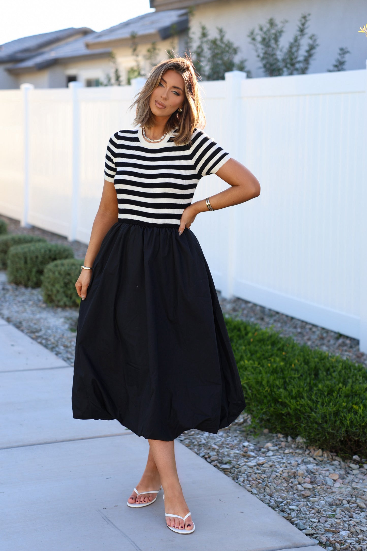 A woman stands outdoors in the Black Stripe Bubble Hem Midi Dress, with a white fence and greenery in the background.