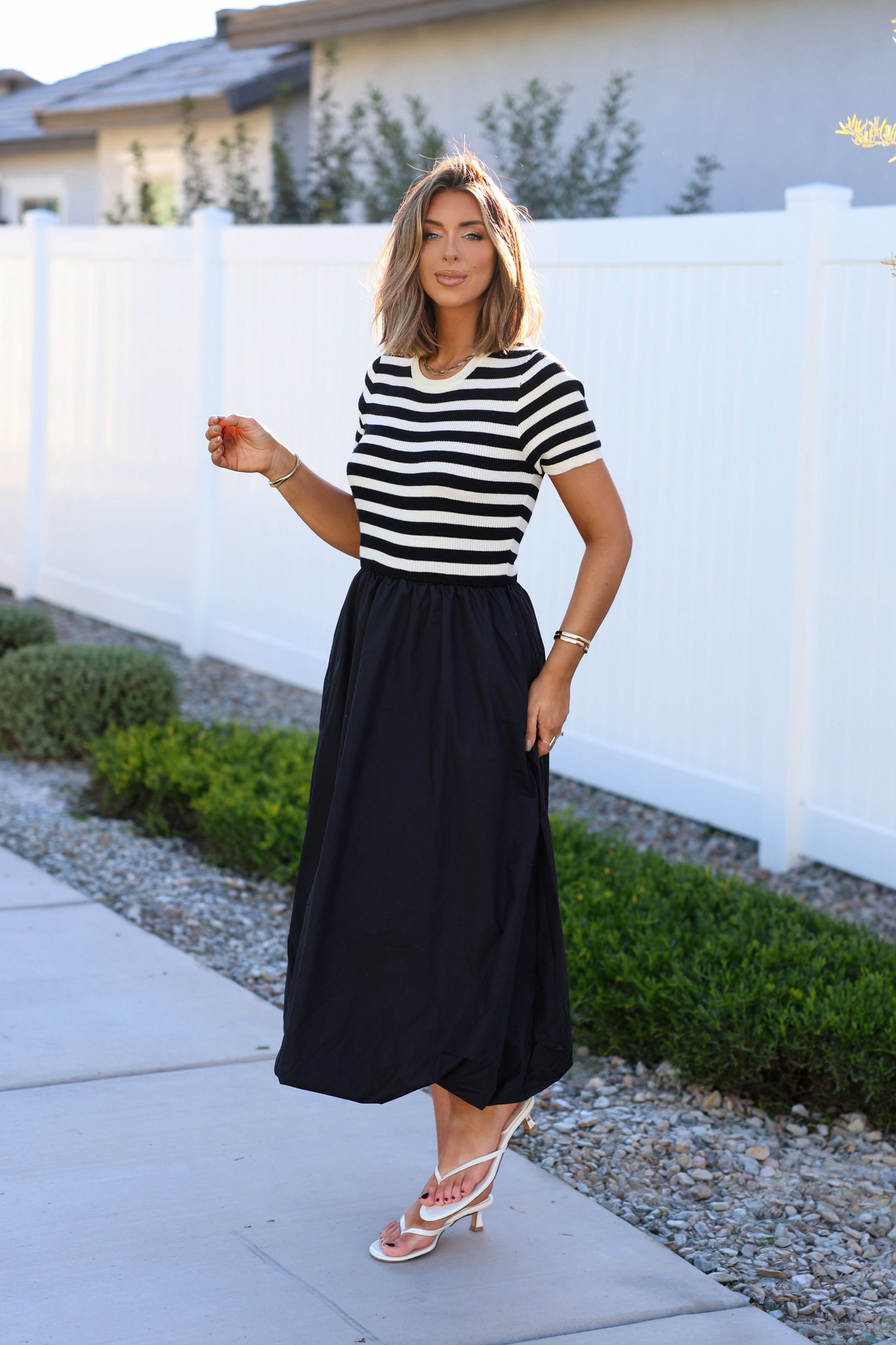 A woman wearing a Black Stripe Bubble Hem Midi Dress stands on a sidewalk by a white fence with greenery.
