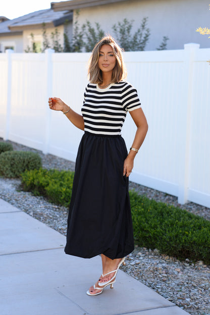 A woman wearing a Black Stripe Bubble Hem Midi Dress stands on a sidewalk by a white fence with greenery.