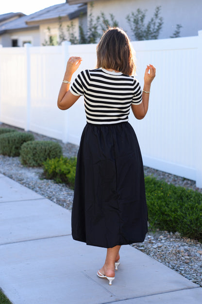 Woman wearing a Black Stripe Bubble Hem Midi Dress walks on a sidewalk by a white fence and greenery, facing away from the camera.