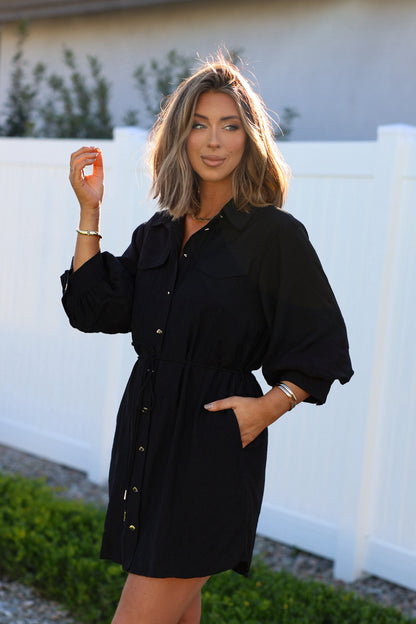 A woman wearing the Black Tie Mini Shirt Dress with an adjustable waist stands by a white fence, one hand in her pocket.