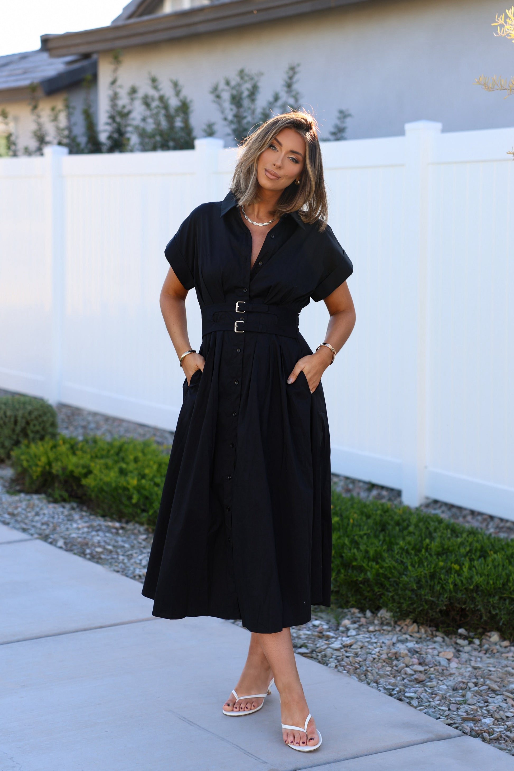 Woman wearing a Black Double Belted Midi Dress stands by a white fence and greenery.