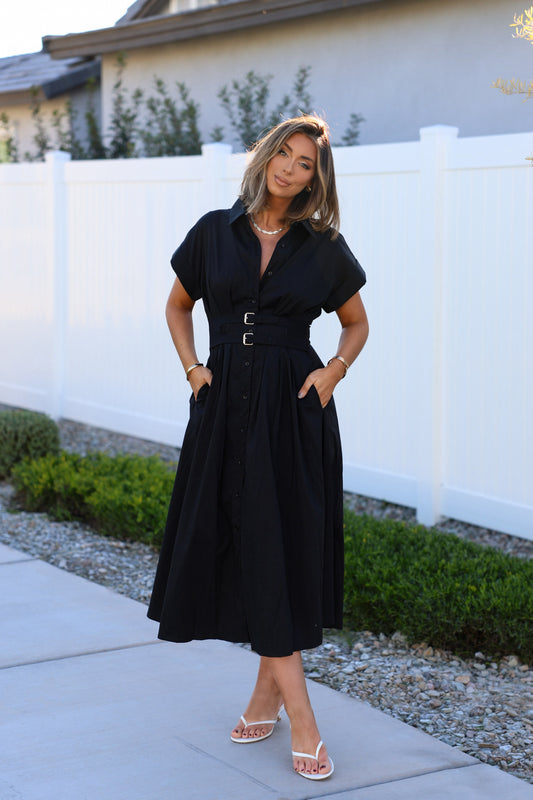 Woman wearing a Black Double Belted Midi Dress stands by a white fence and greenery.
