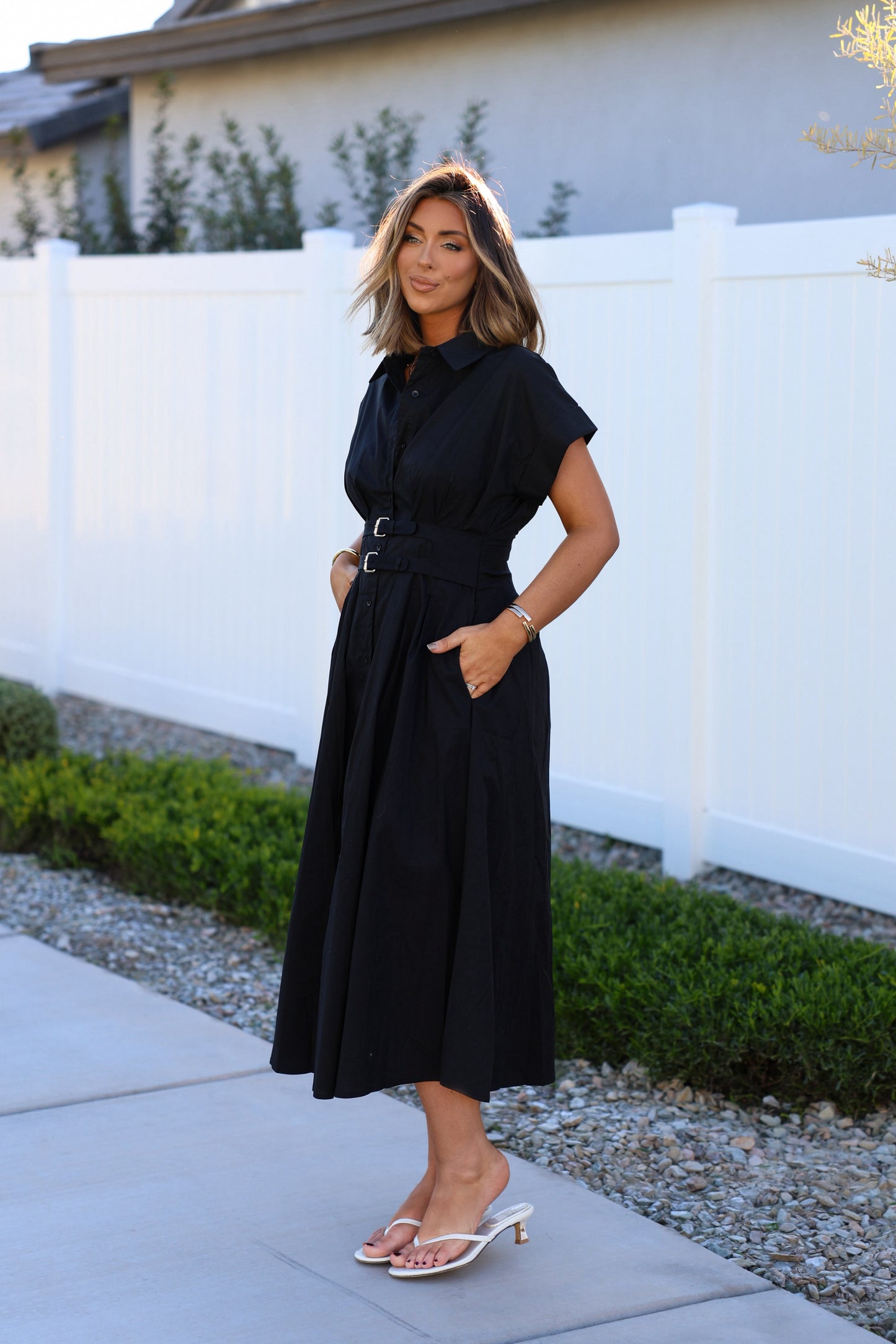 Woman outdoors in a Black Double Belted Midi Dress, posing by a white fence with hands in pockets and wearing heeled sandals.