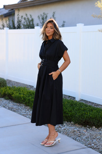 Woman outdoors in a Black Double Belted Midi Dress, posing by a white fence with hands in pockets and wearing heeled sandals.