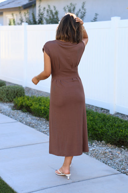 A woman in the Elevated Mocha Fitted Midi Dress and white sandals stands by a white fence, showcasing a minimalist silhouette.