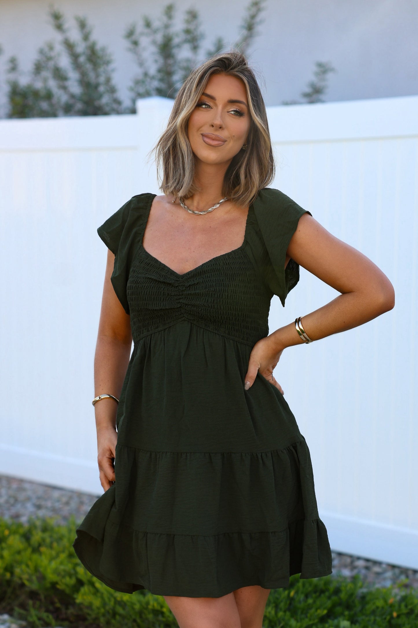 A woman in Ava's Sweetheart Olive Mini Dress stands by a white fence, smiling with one hand on her hip.