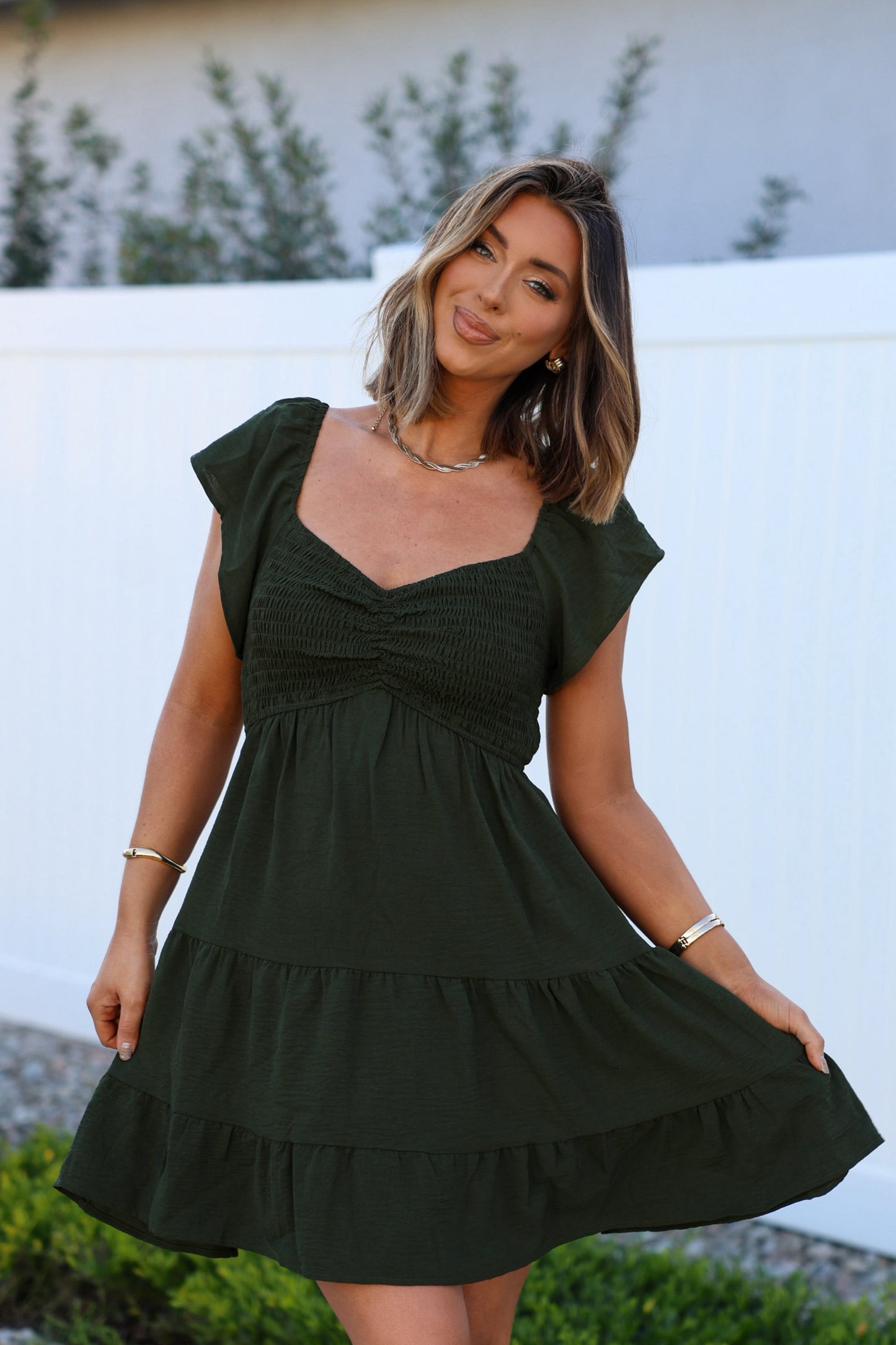 Woman smiles outdoors in Ava's Sweetheart Olive Mini Dress, posing by a white fence with greenery.