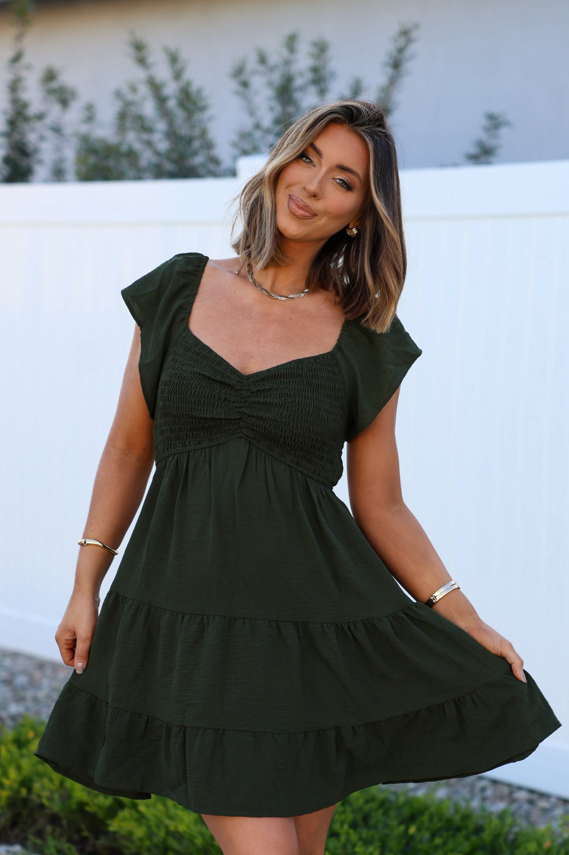 Woman smiles outdoors in Ava's Sweetheart Olive Mini Dress, posing by a white fence with greenery.