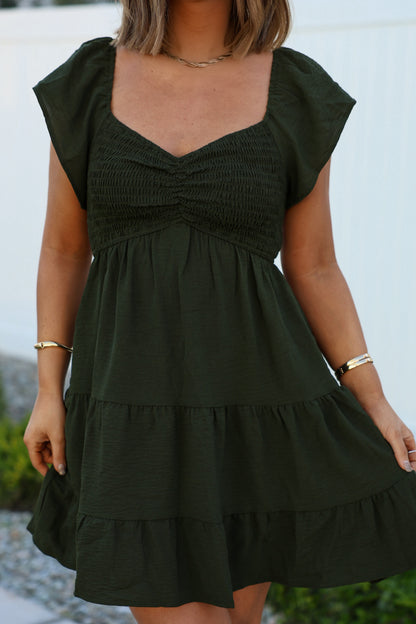 Woman in Ava’s Sweetheart Olive Mini Dress with short sleeves and tiers, standing outdoors against a light background.