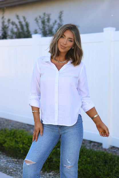 Woman with shoulder-length hair in a Thread and Supply White Button Down Presley Top, standing outdoors by a white fence.