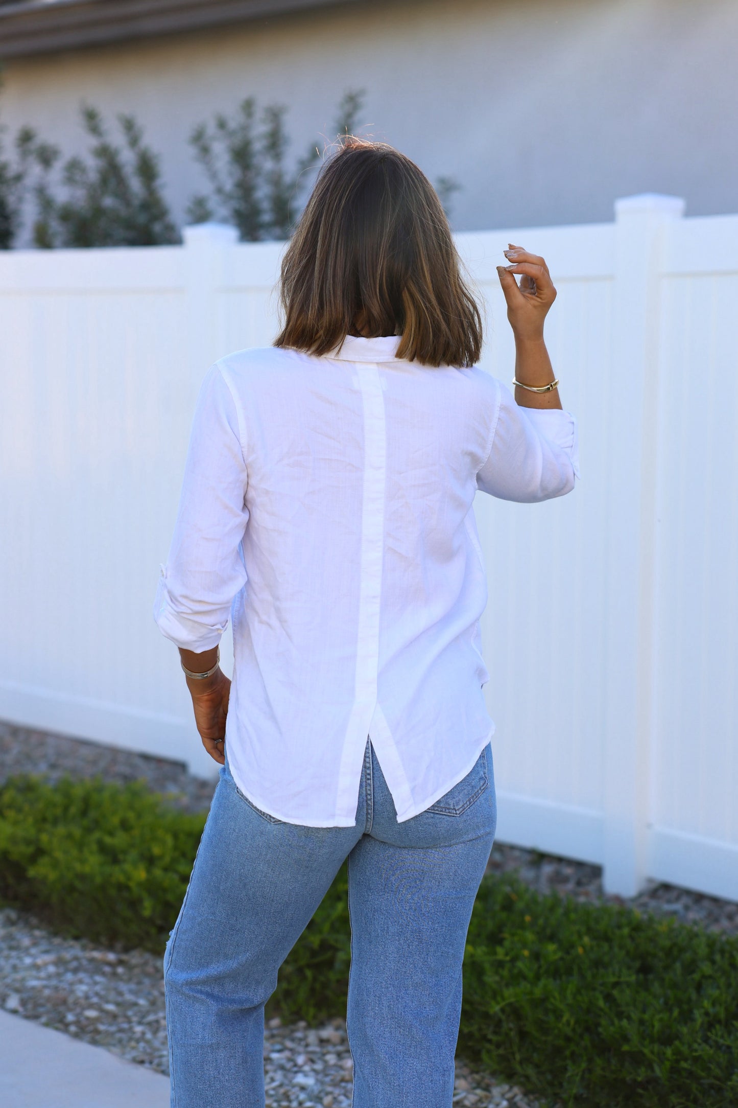 A woman with shoulder-length hair wears a Thread and Supply White Button Down Presley Top and blue jeans by a white fence.