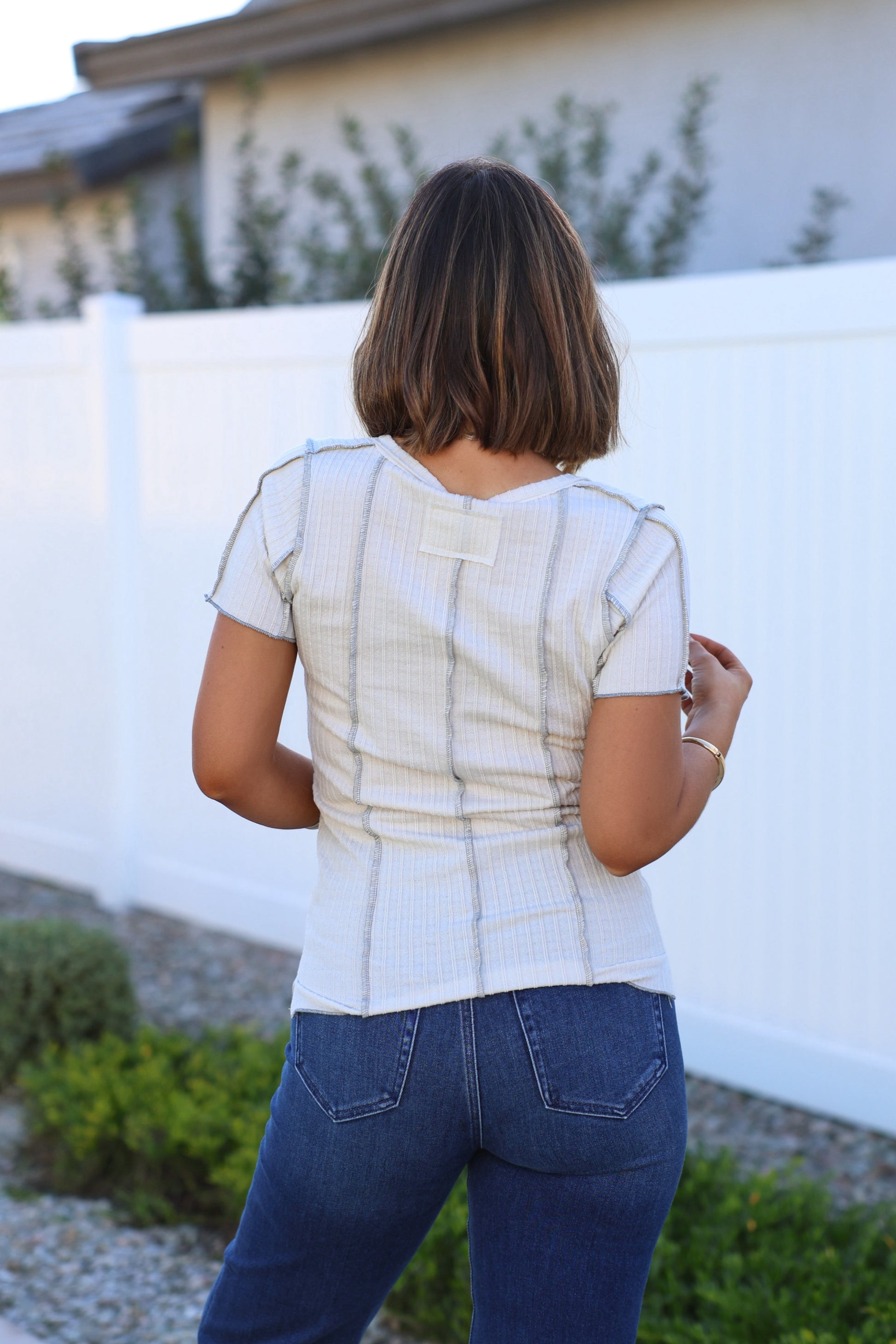 A woman with short brown hair stands outdoors near a white fence, wearing the Natural Contrast Trim Button Up Top and blue jeans.