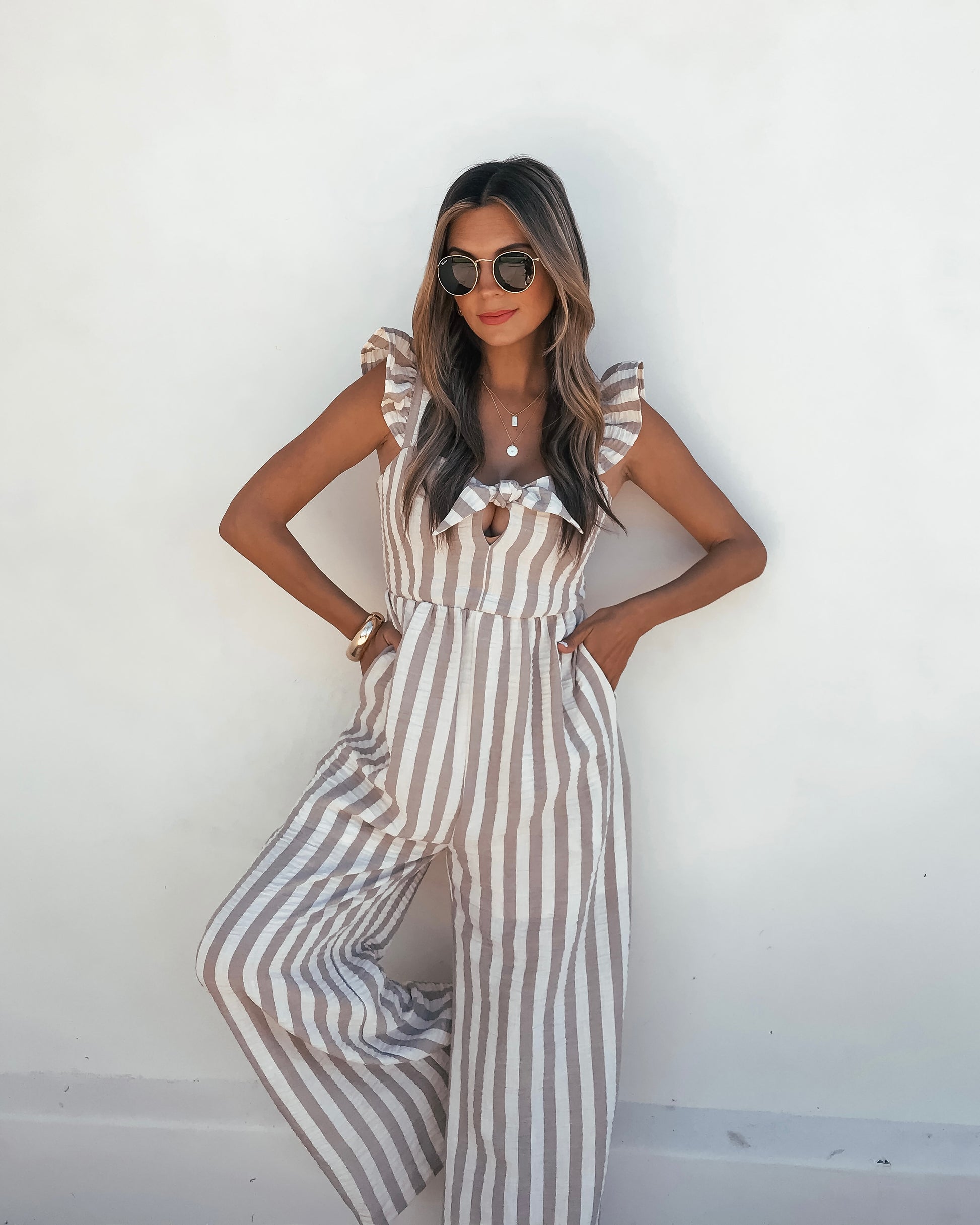 Woman in a Beige Striped Wide Leg Jumpsuit, sunglasses, and red lipstick stands confidently against a white wall.