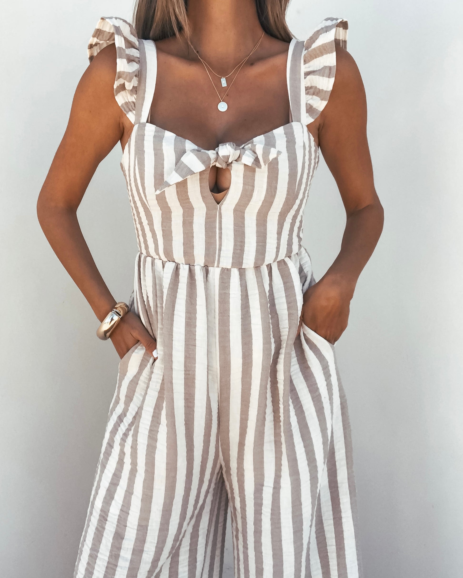 Woman models the Beige Striped Wide Leg Jumpsuit with front knot and pockets, ideal for summer against a plain background.