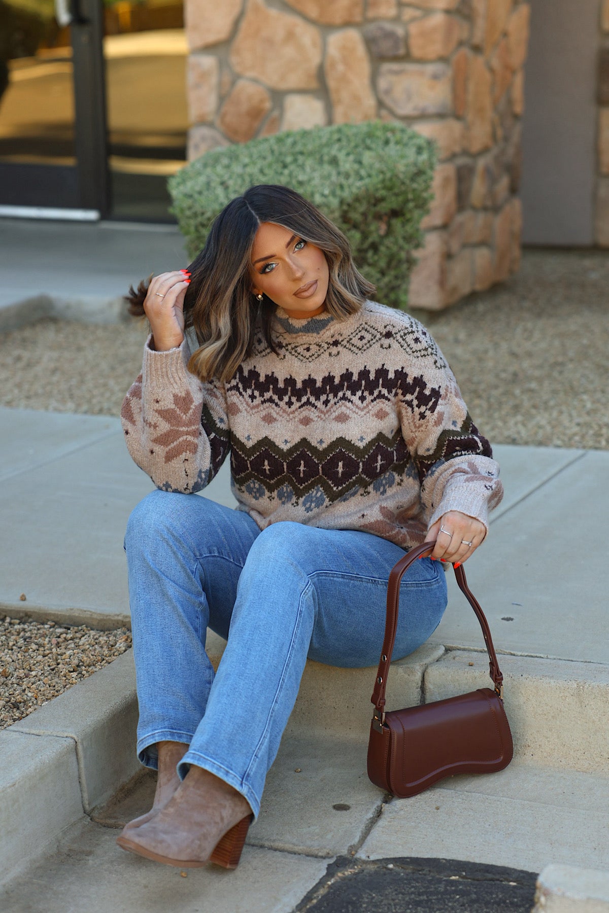 A woman in a Light Khaki Fair Isle Sweater, blue jeans, and ankle boots sits on a concrete step holding a small dark brown handbag.