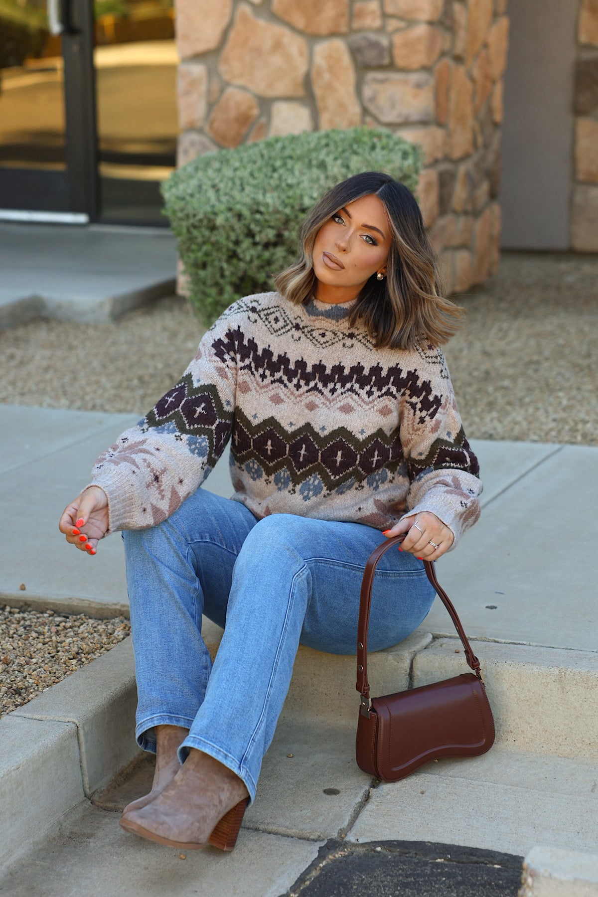 Woman wearing a Light Khaki Fair Isle Sweater and blue jeans, sitting on a concrete step with a brown shoulder bag.