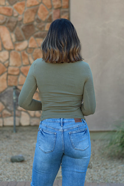 A person in an Ultra Soft Basic Ribbed Top - Dark Olive and blue jeans stands outdoors by a stone wall, with shoulder-length brown hair.