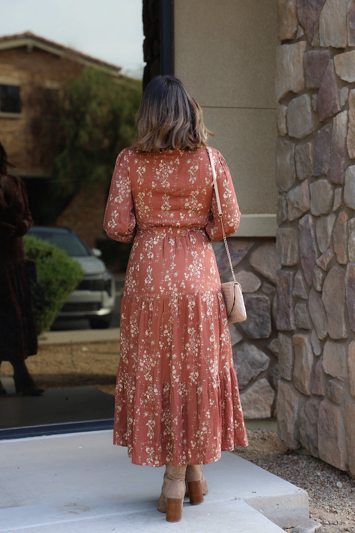 A woman from behind wears a brown Multi Floral V-Neck Midi Dress with a small crossbody bag and tan ankle boots.