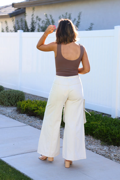 A woman in Cream Wide Leg Drawstring Pants stands outdoors on a sidewalk near a white fence, facing away.