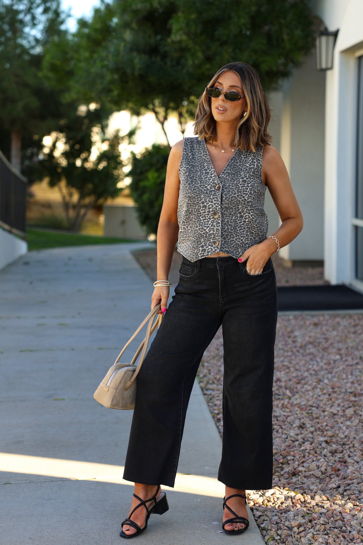 A woman in sunglasses wears a Tan Leopard Button Up Vest, wide-leg black jeans, sandals, and holds a beige handbag outdoors.