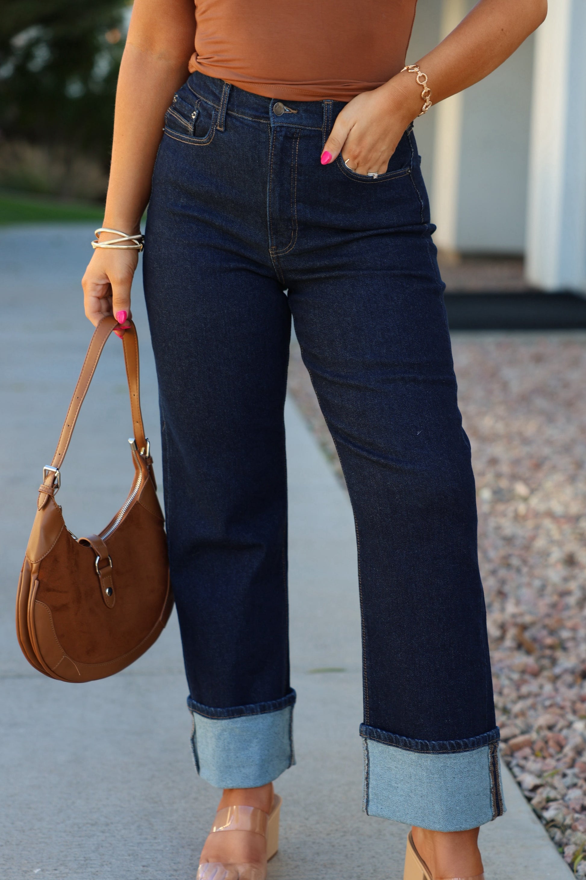 Woman in Dark Wash High Rise Cuffed Dad Jeans, tan top, brown purse, pink nails, clear heels, standing outdoors.
