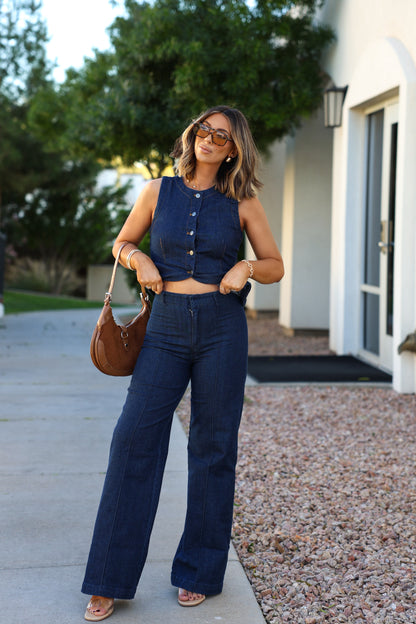 A woman wears a Dark Denim Button Down Vest and wide-leg jeans outside, holding a brown handbag and smiling.