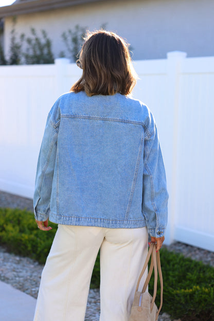 Someone in an Oversized Button Down Denim Jacket stands outdoors with white pants and a tan bag, facing away.