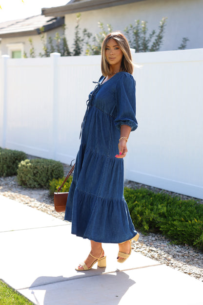 Woman in a Denim Front Tie Tiered Midi Dress and beige heels walks on the sidewalk, carrying a brown handbag.