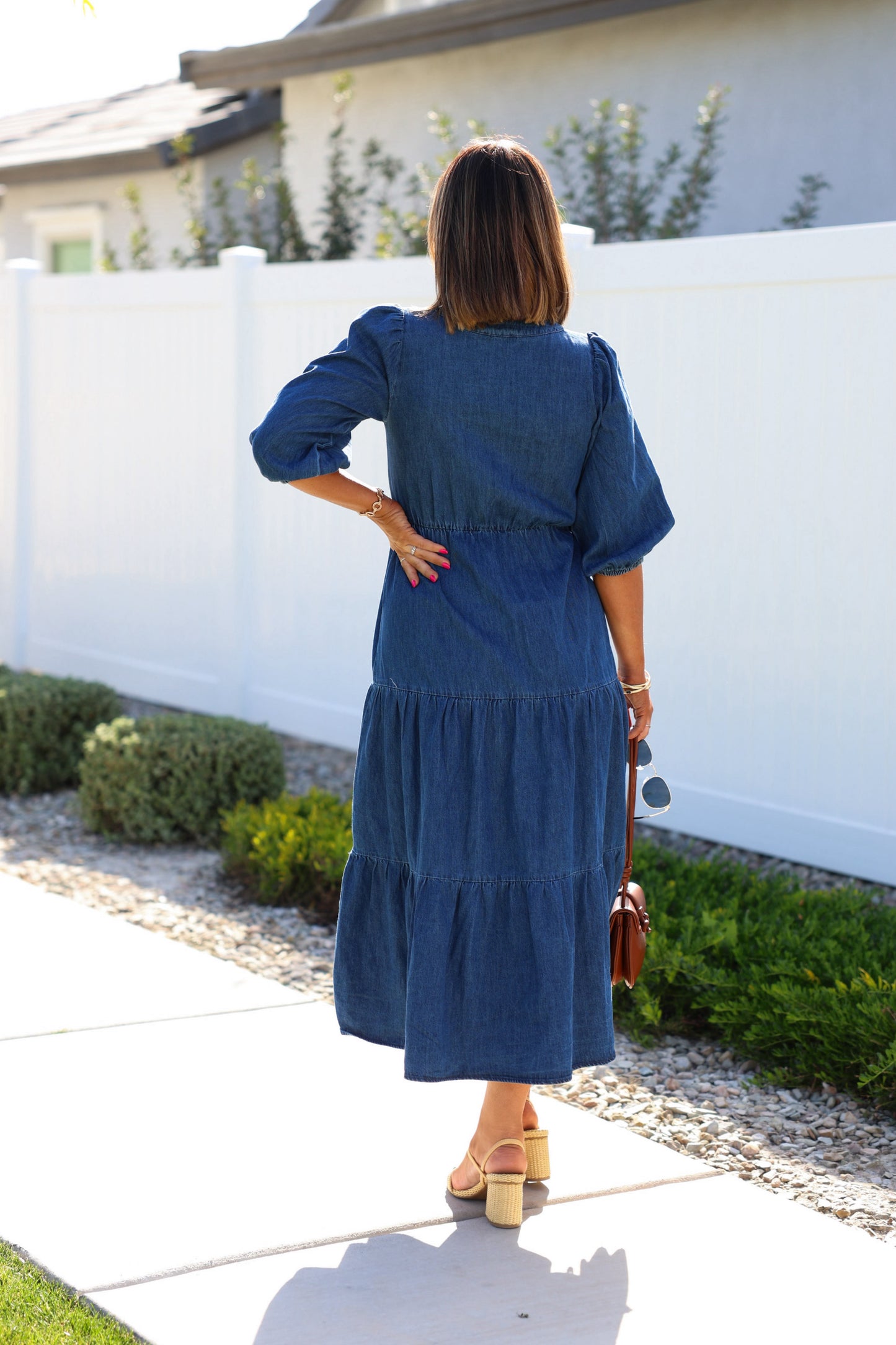 Wearing a Denim Front Tie Tiered Midi Dress and sandals, a woman stands on the sidewalk holding a brown purse and sunglasses.