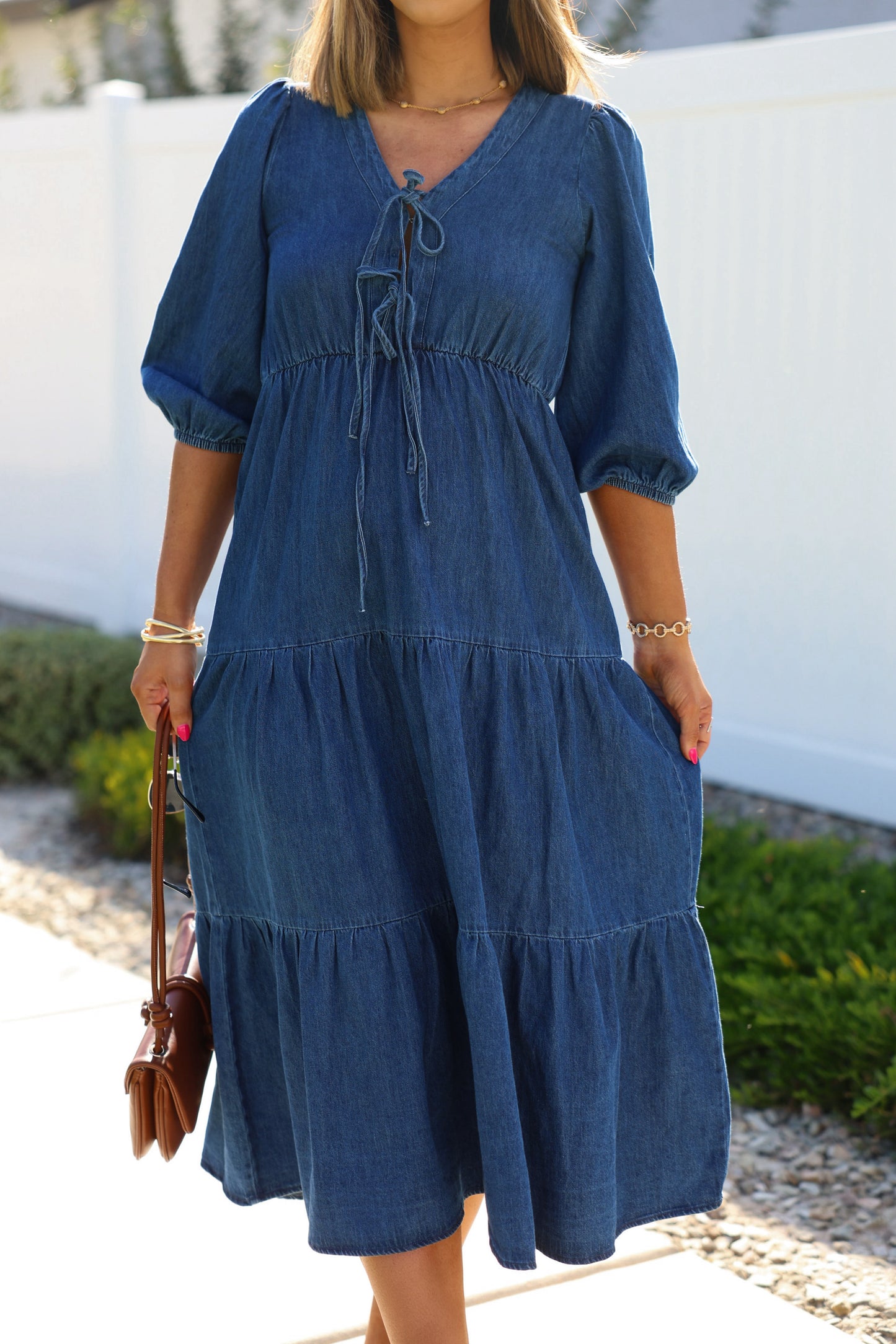 Woman in a Denim Front Tie Tiered Midi Dress, holding a brown purse by greenery and a white fence.
