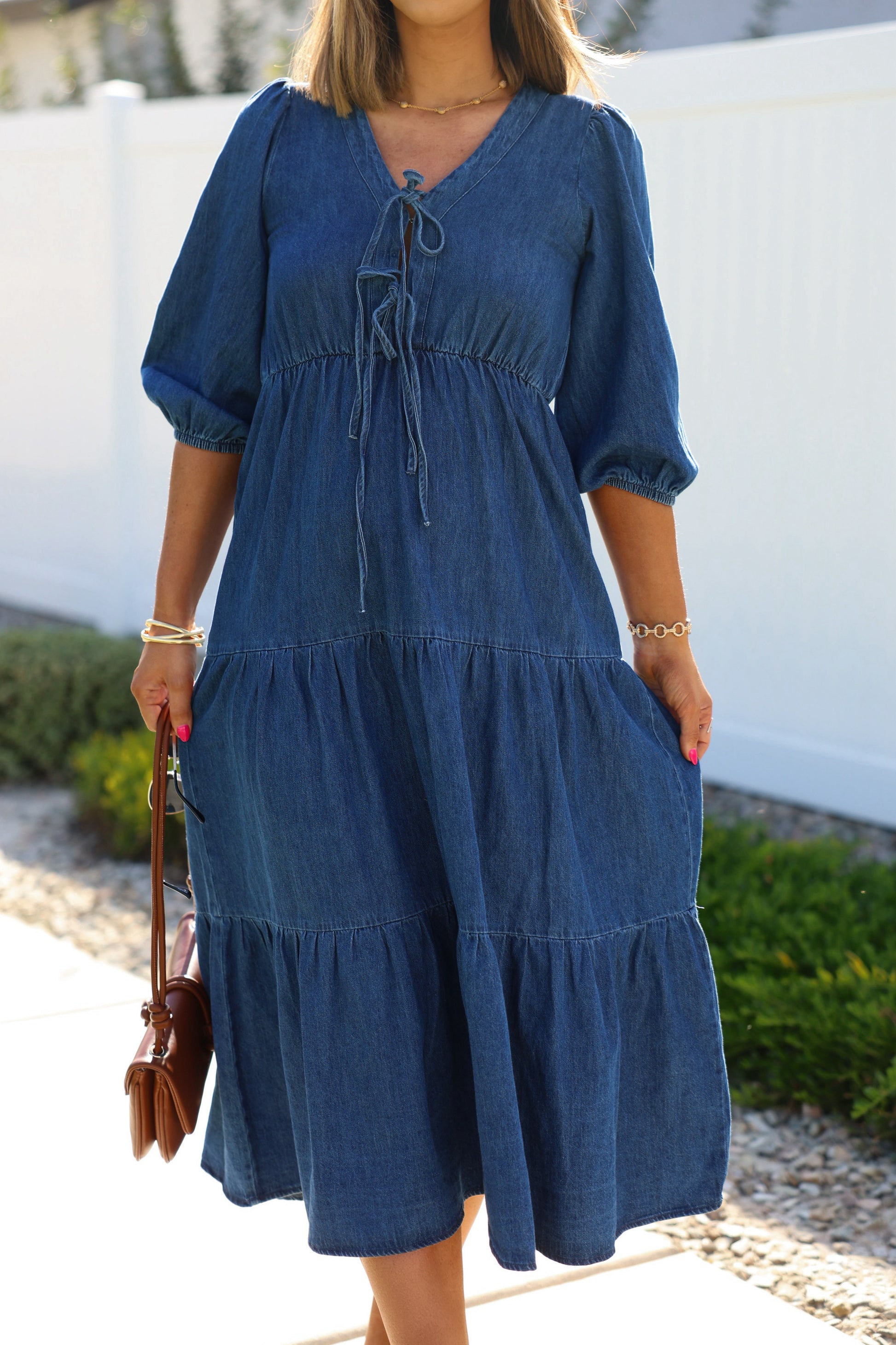 Woman in a Denim Front Tie Tiered Midi Dress, holding a brown purse by greenery and a white fence.
