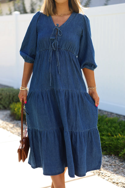 Woman in a Denim Front Tie Tiered Midi Dress, holding a brown purse by greenery and a white fence.