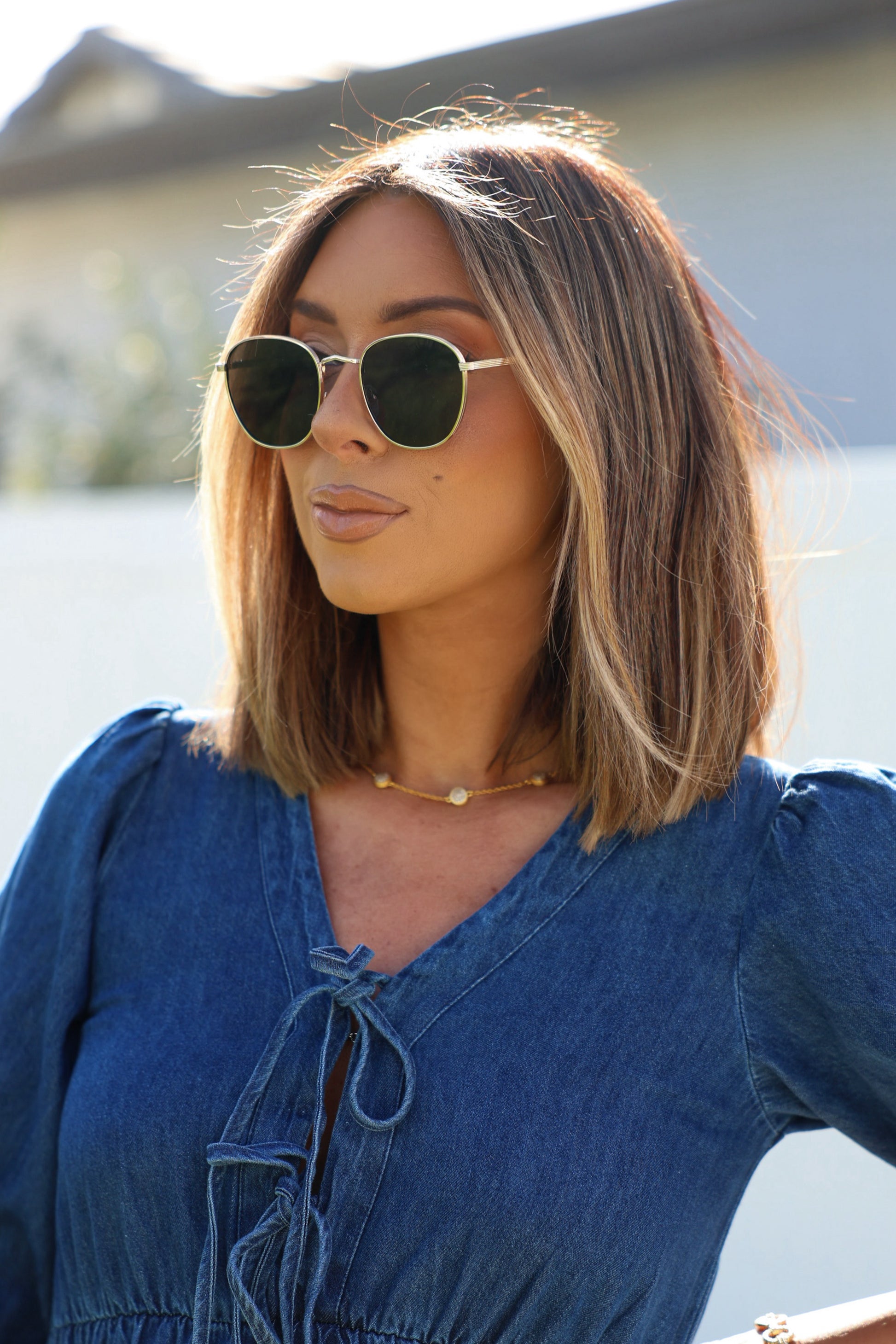 Woman with highlighted hair wears Gold Smoke Lens Round Sunglasses, a denim top, and a delicate necklace outdoors.
